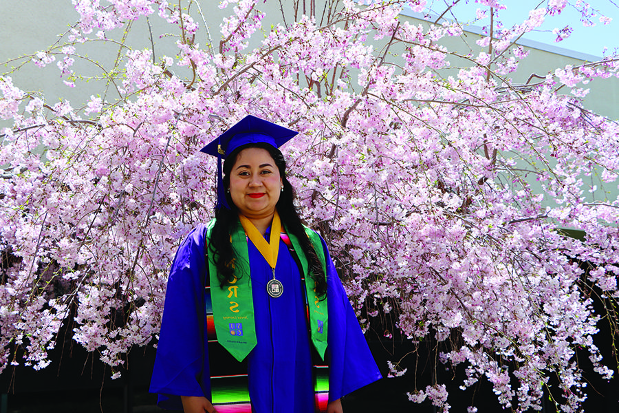 Graduate in full gown with honors tassels in front of pink flowering tree